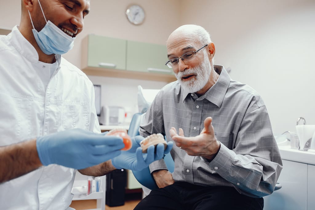 A dentist holding dentures in gloved hands while explaining treatment to an elderly male patient