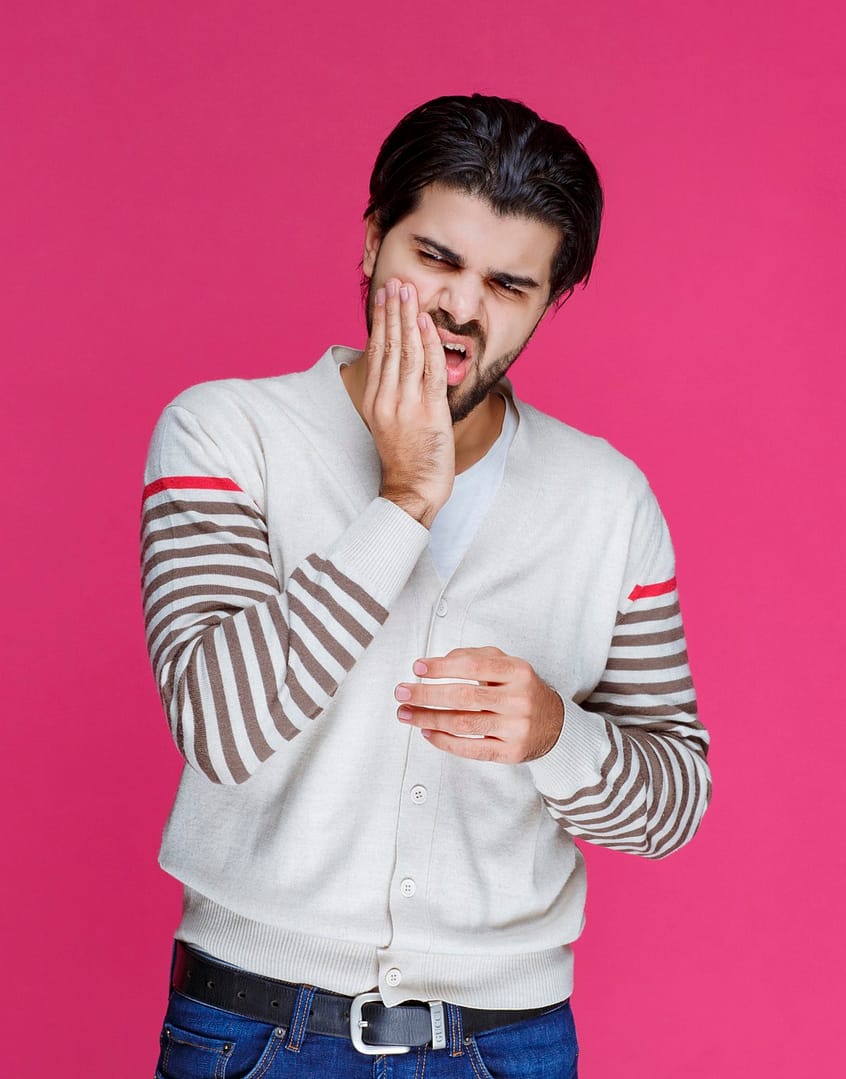 A man standing in front of a pink background holds the side of his face due to tooth pain.