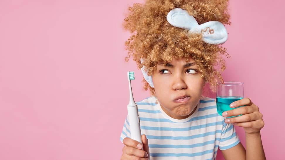 Young woman swishing with mouthrinse while holding an electric toothbrush and looking skeptical